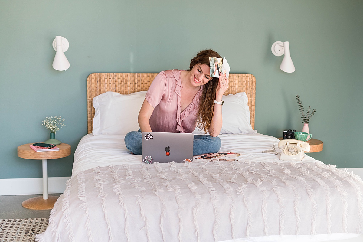 Woman sitting on bed holding photos and looking at laptop